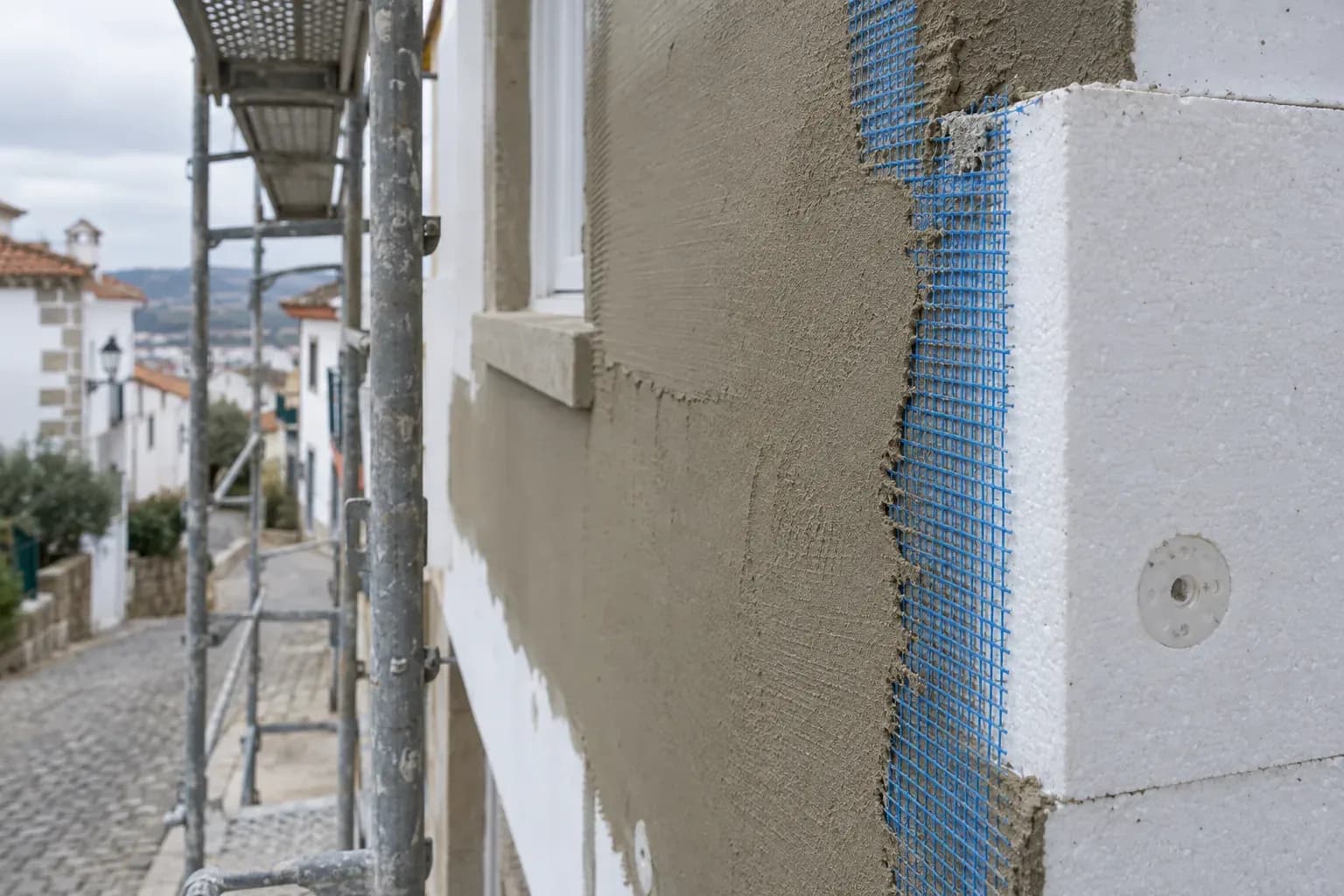 Parede exterior em obra: placa isolante branca fixada com buchas, camada de base e malha azul por acabar.
