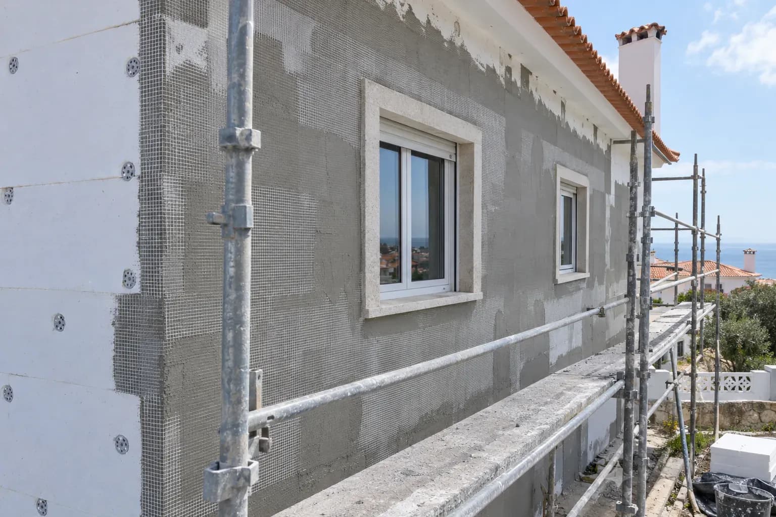 Fachada de moradia com a primeira camada de reboco e rede de malha sobre as placas de isolamento, e andaime.