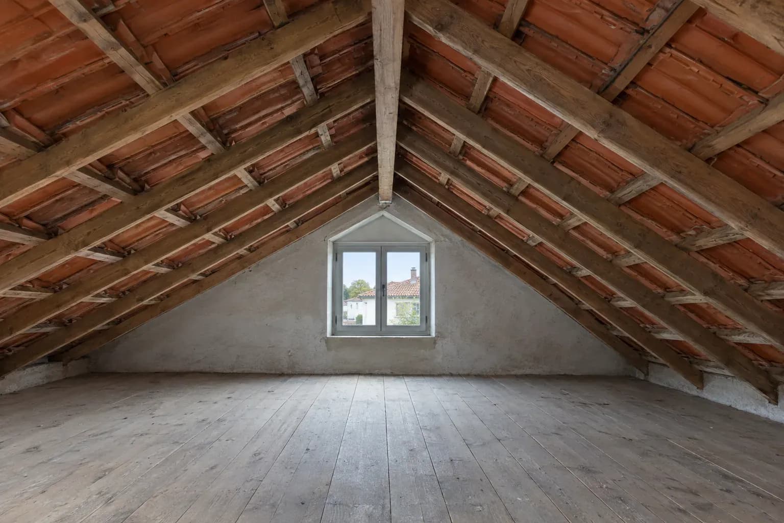Sótão vazio sob o telhado inclinado, com vigas de madeira e telhas à vista, e uma janela pequena